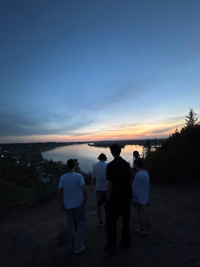 Several people stand on the edge of a cliff overlooking a body of water and a city illuminated by streetlights in the distance, with a sunset in the sky. The photograph was taken in Ольховка, Russia.
