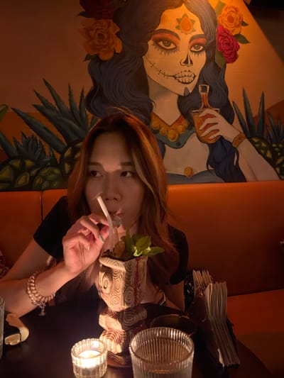 A young East Asian woman is captured in a dimly lit indoor setting, likely a bar or restaurant, during the evening or night. She has long, reddish-brown hair and looks directly at the camera with a neutral expression. Her left hand holds a thin, white, rolled object, possibly a cigarette, near her mouth. On her right wrist, she wears multiple pearl and beaded bracelets.
In front of her, a drink garnished with mint leaves is visible, along with a small, lit candle in a ribbed glass holder, providing warm illumination. Another ribbed glass and a stack of white napkins are on the table. The background features a large, colorful mural depicting a stylized female face, resembling a Calavera Catrina or sugar skull from Día de Muertos imagery. The face has elaborate eye makeup, a rose on its forehead, and a stitched mouth. The figure's dark, wavy hair is adorned with red and yellow roses, and a hand holding a bottle is depicted. Pineapple and agave plant motifs complete the mural, suggesting a Latin American or Mexican theme for the establishment. The visible seating appears to be an orange upholstered booth. This scene takes place in Longta, China. No text is visible in the image.