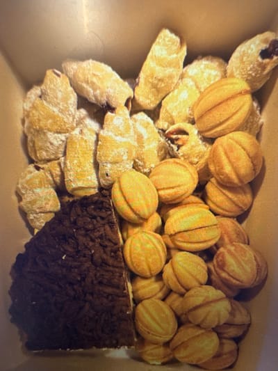 The image displays a close-up, top-down view of a collection of pastries in a light-colored cardboard box. The pastries are varied in appearance. On the left side, a large, dark brown, rectangular piece of cake or brownie is visible, with a textured, slightly crumbly surface. To the right of this, and filling the rest of the box, are numerous small, light brown, oval-shaped cookies or pastries. These cookies have a distinctive ribbed pattern on their surface, resembling halved walnuts or small buns. Some of these cookies are dusted with a white powder, possibly powdered sugar. Several of the cookies appear to be filled with a darker substance, possibly jam or cream. The overall impression is of a confectionery assortment. No specific location in Murcia, Spain is identifiable from the image itself.