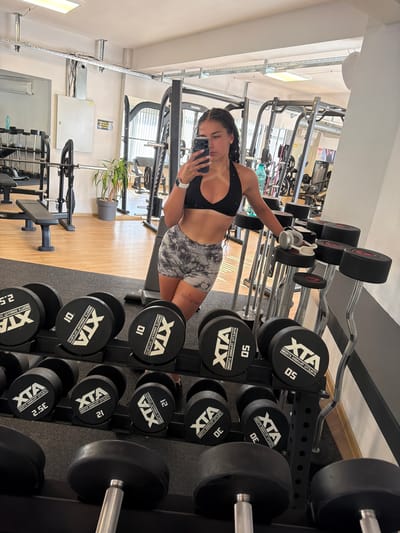 A young woman with dark braided hair is taking a mirror selfie in a gym located in Gabrovo, Bulgaria. She is wearing a black sports bra and grey and white tie-dye athletic shorts. The gym has a polished wooden floor, and various weightlifting equipment is visible, including racks of dumbbells of different weights, weight benches, and squat racks. A potted plant is situated in the background. The lighting appears to be fluorescent, typical of an indoor gym environment. The woman is holding a smartphone in her right hand, and a water bottle is visible on a surface behind her.