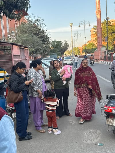 This image depicts a bustling street scene in Jaipur, Rajasthan, India, characterized by reddish-pink architecture. In the foreground, a group of six individuals, primarily women and children, are positioned on the roadside.

From left to right: A person's lower torso in a light blue garment is partially visible. Next, a woman with dark hair in a black quilted jacket and blue jeans holds a brown shoulder bag and observes a mobile phone. Beside her, a blue scooter is partially visible. A second woman, with dark hair tied back, wears a patterned white and black top over a purple salwar kameez and holds a phone to her right ear, appearing to be in conversation. Standing between these two women is a small child, a toddler, seen from behind, wearing a striped brown, white, and black long-sleeved top, brown pants, and white shoes. This child appears to be interacting with the woman in purple, possibly holding hands. To their right, a third woman in an olive green long-sleeved top and black flared pants holds an infant dressed in a pink hooded outfit with ear details. The infant faces away from the camera. Further to the right, an elderly woman with glasses, wearing a dark red patterned sari and a matching shawl, stands on the edge of the asphalt, looking towards the camera.

In the immediate foreground on the right, the rear of a red scooter with chrome handles and a license plate reading "RJ05S 0159" is visible. The asphalt road shows some wear and minor debris.

The background reveals a street with moderate vehicular traffic, including several parked cars (white sedans/SUVs) on the left side and moving vehicles further down the road, including a person on a motorcycle on the far right. Tall ornamental streetlights with multiple lamp heads line the street. Prominent reddish-pink architectural structures, characteristic of Jaipur, are visible on both sides of the street, including a large, ornate pillar-like structure on the right and multi-story buildings on the left. Trees with sparse foliage are interspersed along the street, and the sky is clear and pale blue, indicating daylight, possibly late afternoon. On the far left, a utilitarian metal cage structure, possibly a vendor stall or storage unit, stands next to a building.