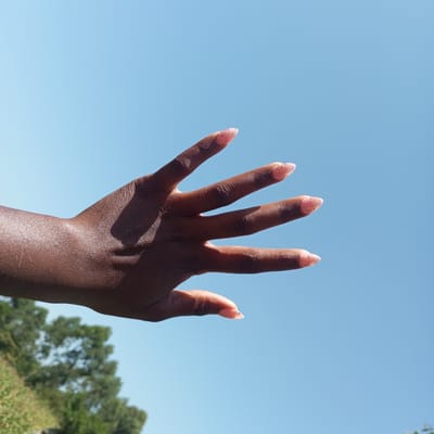 A close-up, low-angle shot shows an outstretched, dark-skinned hand against a clear, pale blue sky. The hand is positioned horizontally in the frame, with fingers splayed and pointing towards the right. The fingers have light pink nail polish with glitter. Sunlight casts a shadow across the palm and wrist. In the lower left corner, out of focus, is a lush green hillside with trees. The image is taken outdoors, likely in a natural setting. No specific landmarks or details indicating the city of Mahiakalo ward, Kenya, are visible.