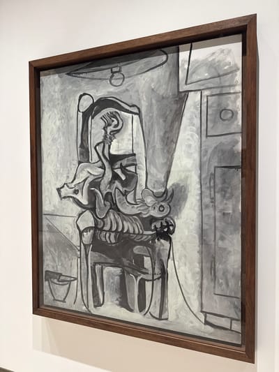 The image shows a framed abstract painting rendered in grayscale, displayed on a white wall. The painting depicts a figure in a chair, with elements that suggest an animalistic form, possibly a bull, due to the presence of horns. The style is cubist, with fragmented planes and distorted perspectives.

The painting is displayed in the Museo Picasso Málaga, located in the Buenavista Palace in Málaga, Spain. The museum houses a significant collection of works by the artist Pablo Picasso, who was born in Málaga. The lighting in the gallery appears to be artificial, casting a soft glow on the artwork.