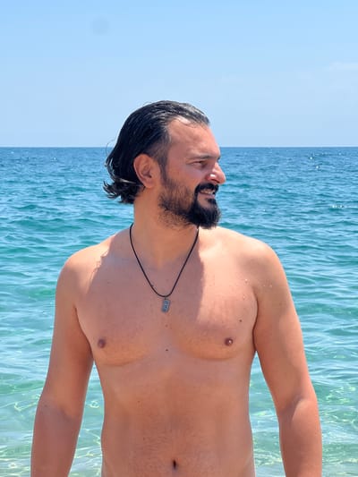 A man with dark hair and a beard stands in the ocean, looking to his right. He is shirtless, wearing a black cord necklace with a rectangular pendant. The water is clear and turquoise, with gentle waves. The sky above is a clear blue. The location is the beach in Sant Pol de Mar, Spain.