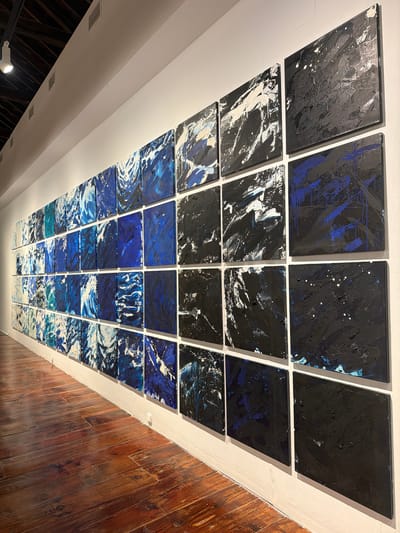 This media file shows a ceiling installation of abstract paintings. The paintings are arranged in a grid-like pattern, with each painting being a square canvas. The paintings are predominantly in shades of black, white, and various blues, with some featuring splashes of color and textured brushstrokes.
The overall impression is one of dynamism and perhaps cosmic or aquatic themes, given the color palette and abstract nature of the art. The paintings are mounted on a white ceiling, with a warm wooden wall visible on the right side of the frame, suggesting an interior space, possibly an art gallery or a modern architectural setting. The lighting appears to be artificial, illuminating the artwork from above and casting subtle shadows. There are no people visible in the media file, and the focus is solely on the artwork and its installation. The time of day and weather are not discernible from the image. No visible text is present. The location context is La Cuesta, Spain.