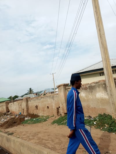 A man walks from left to right across a dirt road in front of a low, weathered stone wall. He is wearing a blue tracksuit with white and red stripes down the sleeves and pant legs. The wall is partly crumbling and has visible holes. Behind the wall, a few buildings with light-colored walls and blue roofs can be seen, along with some trees. The sky is overcast. The road in the foreground is a dirt path with a concrete curb. To the left of the man, there is a pile of debris and garbage. The scene appears to be in an outdoor, semi-urban environment.