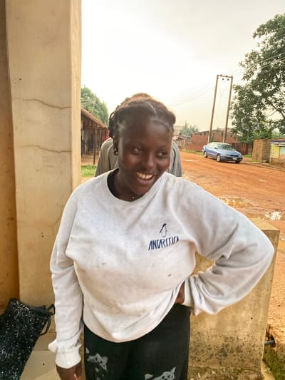 A young woman smiles towards the camera in an outdoor setting in Kuru Dungus, Nigeria. She wears a white sweatshirt with a penguin logo. The background shows a muddy road, buildings, and a blue car. The environment appears rural with basic infrastructure, including power lines.