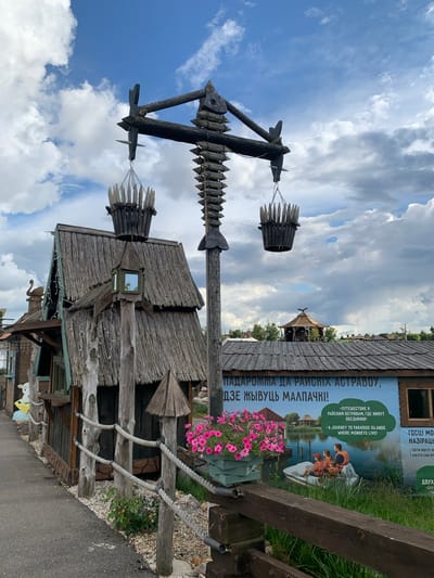 A wooden, rustic-themed building complex is pictured on a cloudy day in Малахавецкі сельскі Савет, Belarus. A dirt path runs along the left side of the frame, bordered by a wooden fence and stones. The main structure features a thatched roof and a prominent wooden post with two suspended wooden buckets. In the background, a large banner depicts a family in a boat on a lake, with text in Belarusian and English. The scene suggests a tourist attraction or recreational area with a naturalistic aesthetic.