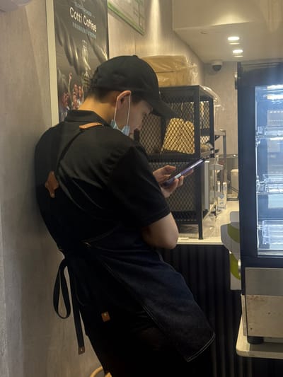 The image shows a person wearing a black cap, a black shirt, and a denim apron, leaning against a wall with a poster for "Cotti Coffee" on it. The person is looking at a mobile phone in their hands. Behind the person, there is a metal shelf with items covered in plastic and a security camera mounted on the ceiling. To the right of the person, there is a display refrigerator with shelves. The environment appears to be a commercial establishment, possibly a cafe or coffee shop, located in Xiangzhou, China.