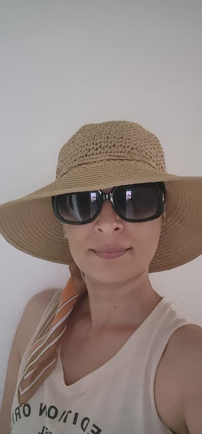 A medium shot shows a woman wearing a straw sun hat and large, dark sunglasses. She is looking directly at the camera, and a faint smile can be seen on her lips. A patterned scarf is visible around her neck, and she is wearing a light-colored tank top. The background is a plain, light-colored wall, suggesting an indoor setting. The image is a selfie, taken from a slightly upward angle. The setting does not provide any indication of being in Sderot, Israel.