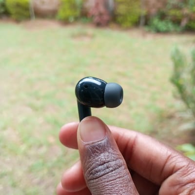 A dark-skinned human hand, primarily the thumb and forefinger, is shown holding a single glossy black wireless earbud. The earbud features a dark grey silicone ear tip and exhibits minor surface abrasions, along with small circular perforations on its body. The background is a heavily blurred outdoor environment, characterized by green grass in the midground and indistinct green and reddish-brown foliage in the distance. The scene is located in Mahiakalo ward, Kenya.