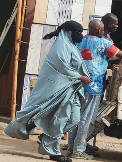 The image shows three individuals in what appears to be an outdoor setting, potentially a marketplace in Maiburiji, Nigeria. The primary subject is wearing a light blue, full-body garment with a dark head covering, positioned in a seated or squatting posture. This individual is partially obscured by the garment they are holding. Behind them, two men are visible, one wearing a blue patterned shirt with red accents and the other wearing a plain grey shirt. The setting includes building materials, potentially tiles, displayed on a rack, and a glimpse of a street or sidewalk.