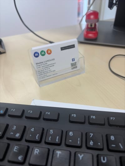 An interior shot depicts a light-colored wooden desk surface. In the mid-ground, a white business card for "Maxime LANGLOIS, Chargé de Clientèle" of "MMA Letellier associés" is displayed upright in a clear plastic holder. The business card includes contact information, a QR code, a Facebook icon, and specifies the address as "3B Bd Maréchal Juin, 06800 CAGNES SUR MER, France." To the right and rear of the business card, a small red object resembling a miniature vehicle or cartoon-like figure, potentially a stapler, rests next to the black base of a monitor stand. Black cables are visible across the desk surface. The foreground is occupied by the top section of a black computer keyboard with white legends, revealing function keys (F2-F7) and number keys (3-8). The background consists of a plain white wall.