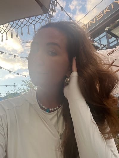 A medium close-up shot shows a person with long, reddish-brown hair and light skin, looking towards the left. Their right hand is touching their ear, with the fingers partially obscuring their face. The person is wearing a white hooded jacket with a visible zipper and a pearl necklace with a blue pendant. The background features a cloudy sky with string lights and a building with the golden lettering "ШИНСКИЙ" visible at the top. The environment appears to be an outdoor seating area of a cafe or restaurant, possibly in Estosadok, Russia.