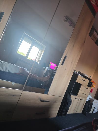The image shows a bedroom interior, possibly in Cherven Bryag, Bulgaria, reflected in a mirrored wardrobe. The wardrobe has light wood-grain doors. The reflection shows a window with bright daylight coming through, indicating daytime. Reflected in the mirror is also a person lying on a bed with their legs raised. A book with a pink cover is visible. Next to the wardrobe is a shelving unit with black and wood panels, topped with personal items and what looks like backpacks. A red Nike shoebox is visible on the top shelf. The background is blurry and dark.