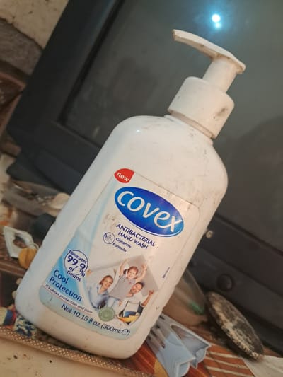 The image shows a white plastic pump bottle of Covex Antibacterial Hand Wash. The bottle is positioned in the foreground, at an angle, and appears to be dirty with smudges and dust. The label on the bottle is mostly visible, indicating it is a "new" product, featuring the brand name "covex" in blue, and details such as "ANTIBACTERIAL HAND WASH", "Glycerine Formula", "*Eliminates 99.9% of Germs", and "Cool Protection". There are also images of a family on the label. The background is out of focus but appears to be an indoor environment with a dark, possibly electronic device, and various objects, including what looks like discarded items. The scene is set in Chobruchi, Moldova.