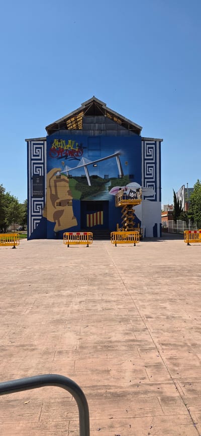 The image shows the exterior of the Trinquet Comarcal building in Sagunto, Spain. The building's facade is painted with a large mural depicting various elements, including a bridge, the Valencian flag, and graffiti. A yellow mobile crane is positioned in front of the building, possibly for maintenance or mural work. Temporary yellow and red barriers surround the building on the paved ground. The sky is clear blue, and trees are visible in the background. A metal railing is in the bottom-right corner of the image.