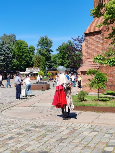 This media file captures a sunny day in a cobblestone plaza, likely in Kaliningrad, Russia. The scene is bustling with activity. Several people are walking around, some appearing to be tourists with luggage, while others are engaged in conversation. In the foreground, a person in a distinctive costume stands facing away from the camera. They wear a red skirt over black pants, a light-colored hooded cloak, and a large, metallic helmet or headdress. The costume suggests a reenactment, a festival, or a street performance.

Behind the costumed individual, a miniature architectural model of a city is displayed on a low platform. Beyond this, a row of buildings with awnings suggests a commercial area with cafes or shops, one of which has Cyrillic text that appears to read "КУХНЯ ДЕ ПРУСС" (Kukhnya de Pruss), implying a "Prussian Kitchen" or restaurant. Further in the background, a colorful pedal-powered vehicle or cart is visible, adding to the lively atmosphere. The architecture of a red brick building, possibly a church or historical structure, dominates the right side of the frame, with its tiled roof and stone foundation. Lush green trees are scattered throughout the plaza, providing shade and contrasting with the bright blue sky. The time of day is clearly daytime, indicated by the strong sunlight and shadows. The overall mood is cheerful and vibrant, typical of a warm, pleasant day in a public outdoor space.
