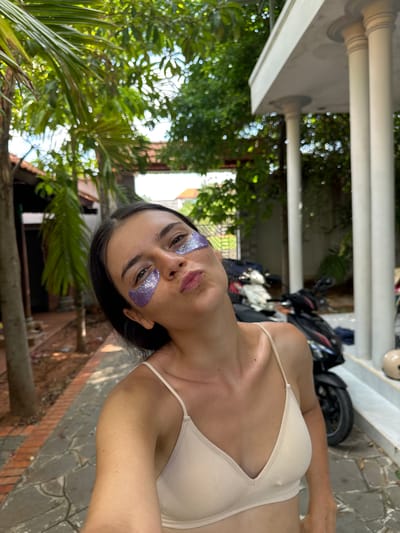 A young woman with dark hair and light skin is taking a selfie outdoors in Mũi Né, Vietnam. She is wearing a beige camisole and has purple glittery eye patches under her eyes. She is puckering her lips and tilting her head slightly. Behind her, a paved walkway leads to a building with white columns. A motorcycle is parked to the right of the woman. Lush green foliage and trees are visible in the background, suggesting a tropical or subtropical environment.