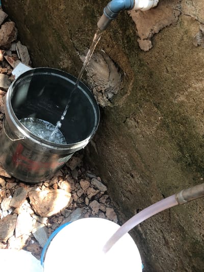 In the city of Alor, Nigeria, an outdoor scene depicts water collection from two distinct sources. In the upper left quadrant, clear water flows from a grey pipe elbow, connected to a white pipe, into a dark-colored metal bucket positioned below. This bucket, marked with red characters on its side, is partially filled with water. To the lower right, a translucent hose, originating from an unseen source, extends into a white bucket with a blue rim, also indicating water collection. The immediate ground surface is composed of scattered concrete debris and rocks. A textured, weathered concrete wall, exhibiting patches of darker discoloration, forms the background on the right side of the image. The scene is illuminated by natural light, casting subtle shadows among the debris. No individuals are present in the frame.
