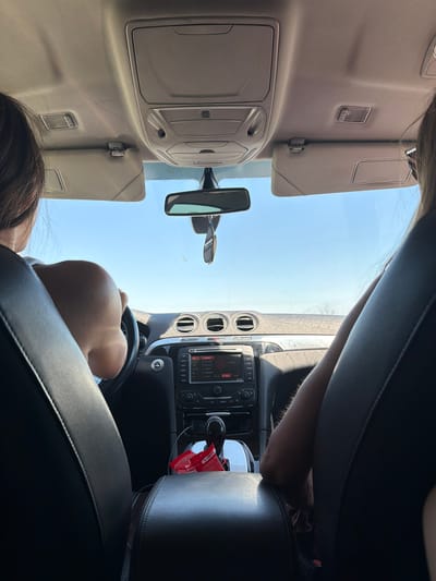 The image depicts the interior of a car, viewed from the back seat. Two individuals are present, one driving and another in the passenger seat. The driver's hands are on the steering wheel, and their arm is resting on the center console. The passenger's arm is visible on their lap. The car's dashboard features a navigation system and air vents. A car air freshener hangs from the rearview mirror. The scene suggests a journey through a rural or open landscape, possibly on a highway, given the clear blue sky and distant horizon visible through the windshield. The specific location of Dinkata, Bulgaria, is not discernible from the visual information in the image.