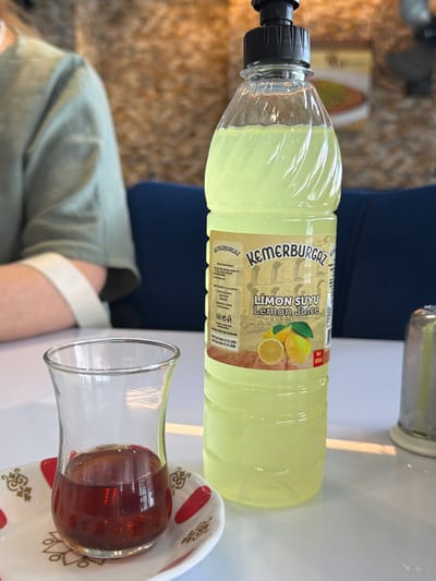 A close-up shot captures a table setting in Beyoğlu, Turkey, featuring a glass of dark amber liquid, likely tea, sitting on a decorative saucer. Beside it, a partially filled plastic bottle of "KEMERBURGÄZ Lemon Juice" stands upright, its label displaying images of lemons and text in Turkish and English. The bottle has a black cap. In the background, a person wearing a light green shirt is partially visible, with their arm resting on the table. The overall environment suggests an indoor setting with a textured wall and a glimpse of a blue upholstered seat. The lighting indicates a daytime scene.
