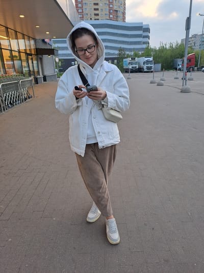 A young woman stands in a public outdoor area, likely a sidewalk or paved walkway, within the city of Elektrostal, Russia. She is dressed in light-colored clothing, including a white hooded jacket, brown trousers, and white sneakers. She is wearing glasses and appears to be focused on a mobile phone or device she is holding in her hands. 

Behind her, there is the facade of a commercial building, possibly a store or supermarket, with a row of shopping carts nearby. In the background, buildings characteristic of a cityscape are visible. Additional elements include street lighting poles, parked vehicles, and greenery. The image appears to have been taken during daylight hours with an overcast sky.