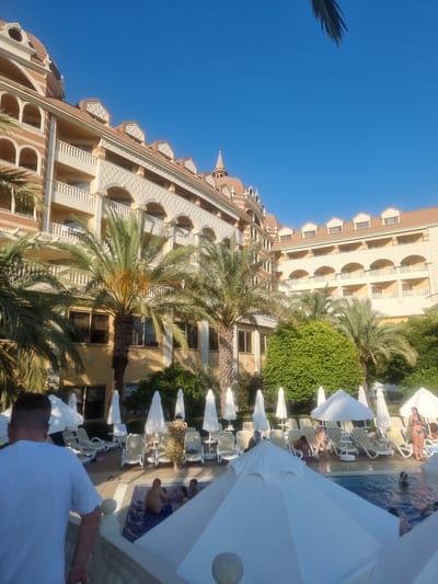 An outdoor resort scene unfolds under a clear blue sky. A multi-story hotel building with ornate architecture, featuring arched windows and balconies, dominates the background. Palm trees are interspersed throughout the property, some bearing fruit. In the foreground, a swimming pool area is visible, with lounge chairs and white umbrellas set up. Several people are relaxing by the pool, with some individuals swimming and others sunbathing. A man in a white t-shirt stands in the foreground, facing away from the camera and looking towards the pool area. The scene appears to be in Manavgat, Turkey, suggested by the architectural style and vegetation typical of the region's resorts.