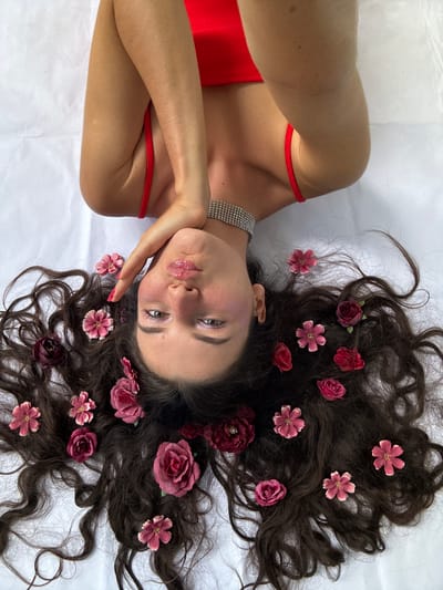 In this media file, a young woman is lying on her back with her head slightly turned towards the camera. Her long, dark, wavy hair is spread out around her head and shoulders, adorned with several artificial flowers in shades of pink and dark red. She is wearing a vibrant red top, possibly a swimsuit or dress, with thin straps. A shimmering, multi-row rhinestone choker is visible around her neck, and a thin red bracelet is on her wrist.
The woman has her left hand resting on her cheek, and her eyes are looking towards the camera. Her lips are pursed, and she appears to be pouting or making a playful expression. Her makeup includes shimmery eyeshadow and a glossy lip color. The overall impression is one of youthful playfulness and a touch of glamour.
The setting is a bright, softly lit environment, suggested by the white background which appears to be fabric, possibly a bedsheet or a studio backdrop. The lighting is even and diffused, casting minimal shadows, which indicates an indoor setting or an overcast day if outdoors. There are no explicit cues in the image to suggest a specific time of day or weather conditions beyond the soft lighting. The location context of Mũi Né, Vietnam, is not visually evident within the image itself, as there are no distinctive landmarks, vegetation, or architectural elements present.
The activity is a posed portrait or selfie, with the woman arranging herself and the flowers for the photograph. The mood is lighthearted and whimsical, enhanced by the floral accents and her playful facial expression. There is no visible text in the media file.