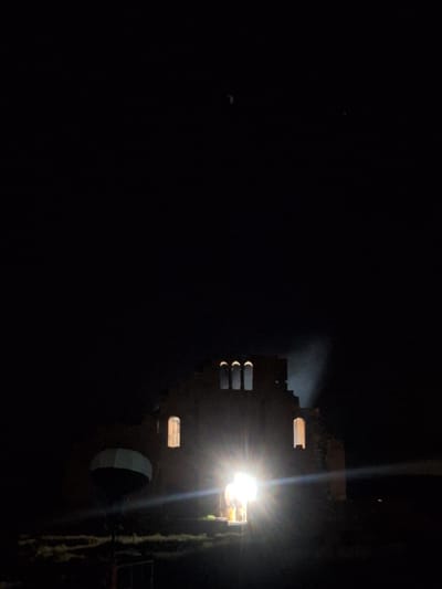 A nighttime image shows a building in Anipemza, Armenia, illuminated by a bright light source. The building appears to be constructed of stone or brick, with several windows and arched openings visible. The overall scene is dark, with most of the image obscured by shadow. The right side of the building is covered in a long strand of foliage, appearing to be vines hanging from the top.