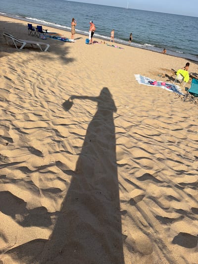 The image shows a beach in Mont-roig del Camp, Spain. The sand is golden and marked with wave-like patterns. Several people are visible on the beach, some walking near the water, others sunbathing or sitting in chairs. Beach chairs and towels are scattered along the shoreline. A long, distinct shadow of a person holding what appears to be a microphone or some similar object stretches across the sand towards the right side of the frame. The sea is calm, and a sailboat is visible in the distance.
