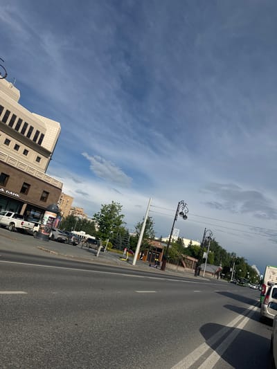 The image shows a street scene in Tyumen, Russia, on a cloudy day with the sun intermittently shining. The perspective is from street level, looking slightly upward towards the sky and across the road.

On the left side of the street, a multi-story building with a light-colored facade and dark rectangular windows dominates the foreground. Below this, a building with a darker brown lower section has signage that reads "А БАНК" (A BANK) in white letters. In front of this building, a white pickup truck is parked. Adjacent to it, a large cylindrical advertising pillar stands.

Further down the street, several cars are visible, including a silver sedan, a dark SUV, and other vehicles further in the distance. Trees and greenery line the sidewalks and are interspersed between buildings. Streetlights with ornate designs are present along the roadside.

The road itself is paved with asphalt and marked with white lane lines. Shadows are cast across the road, indicating the position of the sun. In the distance, more buildings and trees are visible, and a green bus is seen driving down the road. On the right side of the road, a person is visible walking with a yellow electric scooter beside them.