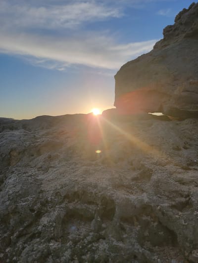 The image captures a low-angle view of a rocky terrain during sunset. The sun, positioned near the horizon, casts a warm glow and creates visible sun flares that extend across the scene. In the background, a clear blue sky is adorned with wispy white clouds. A large rock formation dominates the right side of the frame, partially obscuring the sun. The foreground consists of textured, rough ground covered in stones and rocks. This scene could be located along the coastal cliffs or rocky shores of Quarteira, Portugal, a region known for its natural coastal landscapes.