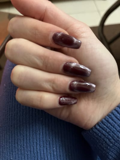 The image displays a close-up of a person's hand, primarily focusing on four manicured fingernails. The nails are painted with a dark, glossy nail polish that appears to be a deep maroon, plum, or burgundy shade, enhanced with a prominent silver or iridescent shimmer. The nails are of medium to long length and have a coffin or tapered square shape. Reflections from a light source are visible on the shiny surface of the polish.

Upon closer inspection, some minor imperfections in the nail polish application or surface texture are noticeable, including slight unevenness or small bumps near the cuticles and along the edges of the nails, and the glitter appears somewhat concentrated in certain areas on some nails. The skin of the hand is fair and appears smooth; no rings or other jewelry are visible. In the upper right and along the right edge of the frame, a section of blue, ribbed, or knitted fabric, likely from a sleeve, is visible, suggesting the person is wearing a sweater or long-sleeved garment. The overall lighting suggests an indoor setting. There is no visible text in the image.