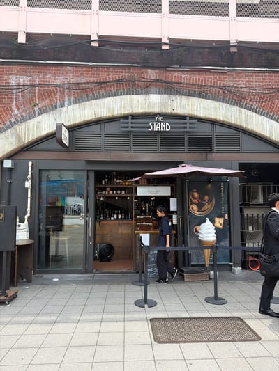 The image shows the exterior of "THE STAND," a shop located in Chiyoda, Japan, likely in a commercial or retail district. The shop is situated beneath a large brick archway, with a pink metal structure visible above. The shop entrance features a glass sliding door and a display area with an outdoor umbrella and a prominent advertisement for soft-serve ice cream. Inside, shelves are stocked with bottles, suggesting it might be a bar or cafe. A young woman wearing a dark uniform is seen exiting the shop, and a man in a suit is walking past on the sidewalk. The sidewalk is paved with grey tiles, and a manhole cover with a circular design is visible in the foreground. The overall impression is of a modern establishment integrated into older architecture.
