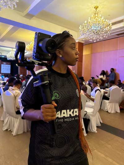 The image depicts an indoor event scene in Itirin, Nigeria. In the foreground, a young Black female, viewed in profile facing right, is carrying a professional camera setup. She wears a dark, patterned short-sleeved shirt with "AAKA CREATIVE MEDIA AG" and a green stylized speaker/microphone logo visible on the front. Her hair is styled in braids, some of which are reddish-orange. She holds the handle of a camera gimbal stabilizer in her right hand, with the mounted dark-colored camera resting on her right shoulder. The camera is equipped with a lens. Her visible ear bears a small, clear stud earring.

In the midground and background, numerous individuals are seated at round tables covered with white tablecloths and chairs adorned with white slipcovers. These attendees are partially visible, engaged in various activities, including looking at mobile devices and conversing. The room features high ceilings from which multiple ornate crystal chandeliers with numerous bulbs are suspended, providing overhead illumination. Walls in the background are light-colored, with sections bathed in purple uplighting. A partial view of a presentation screen or board is visible in the far left background. The overall environment suggests a professional or semi-formal gathering.