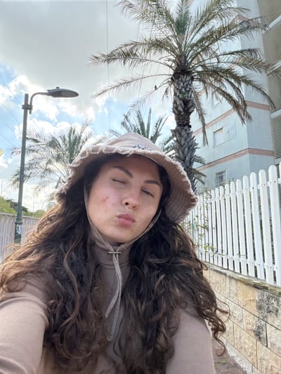 A woman with long, dark brown curly hair is taking a selfie outdoors. She is wearing a light brown hoodie and a matching bucket hat. She has her eyes closed and her lips pursed. Behind her, there's a palm tree, a street lamp, and a building with a white picket fence. The photo was taken in Kiryat Bialik, Israel.