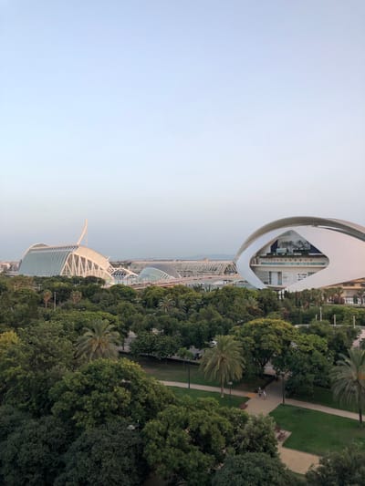 The image captures a panoramic view of the City of Arts and Sciences in Valencia, Spain, bathed in the soft light of dusk. The foreground is dominated by a lush green park with a variety of trees, including palms, and winding pathways. In the midground, several iconic architectural structures of the complex are visible: on the left, the striking L'Hemisfèric with its eye-shaped design, and further to the right, the soaring structure of the Palau de les Arts Reina Sofía. The sky is a pale, hazy blue, indicating the end of the day. A few distant buildings and infrastructure can be seen on the horizon. The scene suggests a tranquil public space, with a couple strolling on a path in the park in the lower right portion of the image.