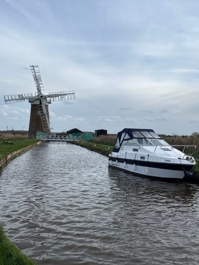 This image depicts Polkey's Mill, a brick tower windmill, located within the Norfolk Broads area of North Norfolk, United Kingdom. The mill, with its four-arm skeleton sails, stands prominently on the left bank of a narrow, rippled waterway, likely a canal or river. A white metal footbridge or walkway extends across the waterway from the mill's base. To the right, a white and dark blue motor cruiser named "Moon Dancer," bearing the registration "706W," is moored parallel to the right bank. The boat has a blue canvas cover over its cockpit area and is secured with white fenders. The banks of the waterway are covered in green grass, with a wooden bench visible on the left bank near the foreground. Beyond the mill, small, dark-colored sheds and areas of tall, dry reeds are visible. The sky is uniformly overcast with light grey clouds. No individuals are present in the scene.