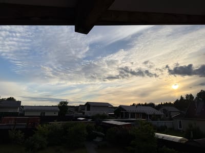 This is a photograph taken from an elevated vantage point, looking out over a suburban or rural landscape under a cloudy sky during what appears to be either sunrise or sunset. The foreground is dominated by a dark wooden beam structure, suggesting the photo was taken from a porch, balcony, or under an overhang.
The mid-ground reveals a collection of houses, varying in architectural style and construction. Some houses appear complete with tiled roofs, while others seem to be under construction, with visible brickwork and exposed framing. Fences delineate property lines, and various outdoor structures, including what look like storage containers and a greenhouse, are visible. Dense greenery, including trees and shrubs, forms a thick layer of foliage throughout the scene, obscuring much of the ground level.
The sky is a prominent feature, filled with a mix of wispy cirrus clouds and darker, more dense formations. The sun is visible as a bright, diffused disc low on the horizon, casting a warm, yellowish light that illuminates parts of the sky and creates a subtle glow. The overall impression is one of a quiet, possibly residential area in Steklyannyi, Russia, during a transitionary period of the day. No people are visible in the image.