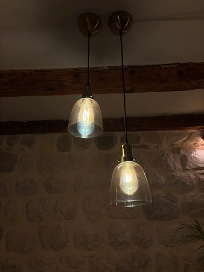 This is a low-angle, close-up photograph of two vintage-style pendant light fixtures hanging from a ceiling in Dobrota, Montenegro. The lights feature clear glass bell-shaped shades enclosing exposed Edison bulbs, each suspended by a black cord from a brass ceiling mount. The fixtures are positioned below a rustic wooden beam that spans the width of the frame. The background consists of a textured stone wall. The lighting is dim, casting a warm glow from the bulbs and creating shadows on the stone wall.