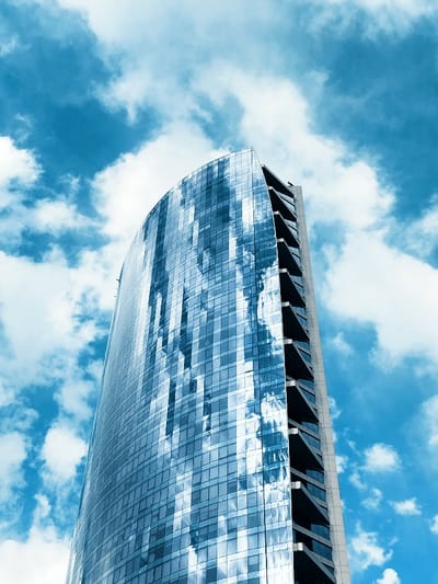 A tall, modern skyscraper dominates the frame, its curved glass facade reflecting the bright blue sky and puffy white clouds. The building's design incorporates a series of stacked, angular balconies on one side. The sky fills the background, contrasting with the building's reflective surface and emphasizing its height and modern architecture. The color palette consists primarily of shades of blue and white.