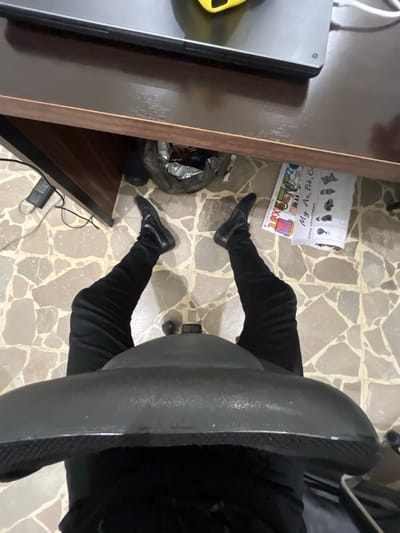 A person sits in a gray office chair, clad in black pants and shoes, with their legs stretched forward. A desk is partially visible on the left, holding a laptop. A plastic bag, filled with items, rests beside the desk. On the floor, a children's alphabet poster lies near the legs. The floor is tiled with irregular beige shapes. Cables run from under the desk.