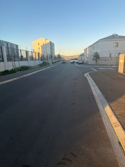 The media file displays an outdoor street scene in what appears to be a modern residential or mixed-use development, likely during either sunrise or sunset. The image is captured from a low angle, looking down a gently curving asphalt road that recedes into the distance.

The street itself is dark asphalt, appearing well-maintained. It is bordered by concrete curbs and gutters on both sides. On the right side, the curb has areas of yellowing or discolored paint. The road shows no significant markings in the foreground but there are faint indications of white lines further down.

Modern, multi-story buildings line both sides of the street. On the right, the buildings are predominantly light-colored (white, beige, or light grey) with multiple windows and some with visible balconies or rooftop structures. One prominent building on the right foreground features a gabled roof and is strongly illuminated by warm, golden sunlight. On the left side of the street, there are also multi-story buildings, some with metal fences or railings along their perimeters. The architecture suggests relatively new construction.

Several vehicles are parked along the left side of the street, including a white sedan and a darker vehicle. More vehicles are visible further down the street on both sides. Small trees and various shrubs are planted along the sidewalks and fences, adding greenery to the urban environment.

The sky is a clear, bright blue, fading slightly to a lighter hue near the horizon. The sun is low in the sky, casting long shadows across the street and highlighting the facades of the buildings it directly illuminates, creating a strong contrast between light and shadow. This warm lighting strongly suggests either early morning or late afternoon.

No people are visible, and there is no direct activity taking place, conveying a sense of quietude. No distinct, legible text is visible within the image. The scene is consistent with a newly developed area in Cape Town, South Africa, as per the provided context.