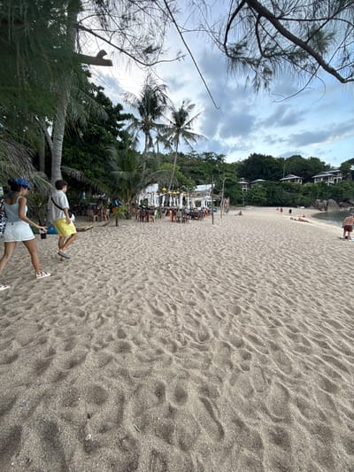 The image depicts a wide sandy beach in Baan Chaweng Noi, Thailand. In the foreground, two individuals are walking away from the camera, one wearing a light blue top and white skirt, the other a white shirt and yellow shorts. The beach is populated with scattered beach chairs and tables, suggesting a dining or lounging area. Palm trees and lush green foliage line the background, with several buildings visible on a hillside further inland. The sky is overcast with clouds. The sand shows footprints and ripples, indicating recent activity.