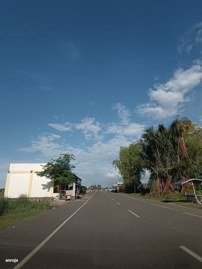 The image captures a road scene in Meunasah Jurong, Indonesia, under a bright blue sky with scattered white clouds. The road is paved and appears to be a two-lane highway. On the left side of the road, there is a white building with a yellow trim and a small tree in front of it, casting a shadow. Adjacent to this building are several structures that appear to be small shops or stalls. On the right side of the road, there are numerous trees, including some palm trees with dried fronds. There is also a small, open-air shelter with a blue tarp. The road stretches into the distance, with a few vehicles visible, including a motorcycle and a car. The overall impression is one of a rural or semi-urban setting in Indonesia, likely during daylight hours. The watermark "anroja" is present in the bottom left corner of the image.