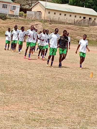 The image shows a group of young men in white and green soccer jerseys, walking in a line on a dry, grassy field in Kipkarren, Kenya. The field is bordered by a brown dirt embankment in the background, with a simple building and a wooden fence.  The men are wearing shorts and are mostly barefoot, but some have sneakers. An orange cone sits at the right edge of the field.  The men are walking toward the camera, their faces showing concentration.