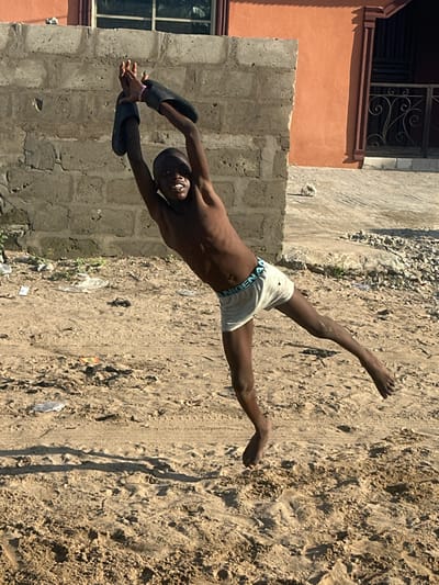 A young boy, likely in his early teens, is captured mid-jump in this photograph. He is shirtless and wearing white boxer briefs with a blue waistband. His body is arched backward, and he is holding a pair of black flip-flops above his head with both hands. His facial expression is one of exertion and perhaps joy. The ground beneath him is sandy and uneven, with some debris and tire tracks visible. To his right, a rough-hewn concrete wall forms the side of a building, painted in places with an orange hue. A window with a grid is visible on the building. The lighting suggests it is daytime, with strong shadows indicating the sun is relatively high. The overall scene depicts a moment of energetic play in an outdoor, somewhat rustic setting, likely a street or yard in Badagry, Nigeria.