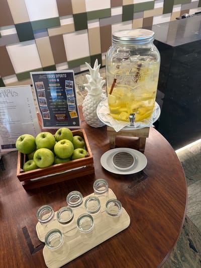 The image depicts a self-service beverage station, likely within a hotel or restaurant. A large glass dispenser filled with ice, lemon slices, and cinnamon sticks, contains a clear liquid, presumably infused water. It is positioned on a wooden stand. Beside it, a white decorative pineapple and a brochure for "JMC TOURS OPORTO" are visible. In the foreground, a wooden crate overflows with fresh green apples. A tray with six small shot glasses is placed on a polished wooden table. The table's surface exhibits wood grain and a decorative inlaid pattern. In the background, a wall with a geometric pattern of squares in beige, green, and white tones is visible. A dark countertop is also present to the right. The scene appears to be set in a contemporary establishment, possibly in Espinho, Portugal, given the tour brochure. No people are present in the image.