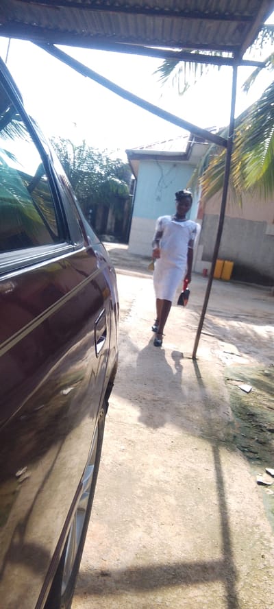 In Madalla, Nigeria, a young Black female, dressed in a white dress and black heels, walks away from a dark maroon car parked on a concrete surface. She carries a black clutch in her right hand and a light-colored object, possibly a brush, in her left. The scene unfolds outdoors, likely in a residential area, with a light blue building and palm trees visible in the background. A corrugated iron roof extends from the upper left of the frame, creating a shaded area. The ground is a mix of concrete and dirt, with some debris scattered around. The sunlight casts distinct shadows of the woman and a vertical pole onto the ground.